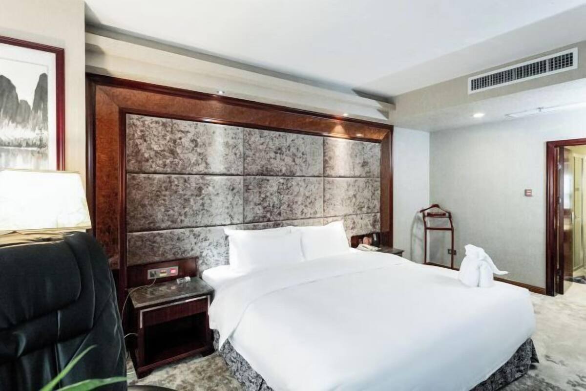 Une chambre d’hôtel avec un grand lit, une tête de lit en bois, une table de chevet, un bureau, une chaise et un tableau encadré au mur.