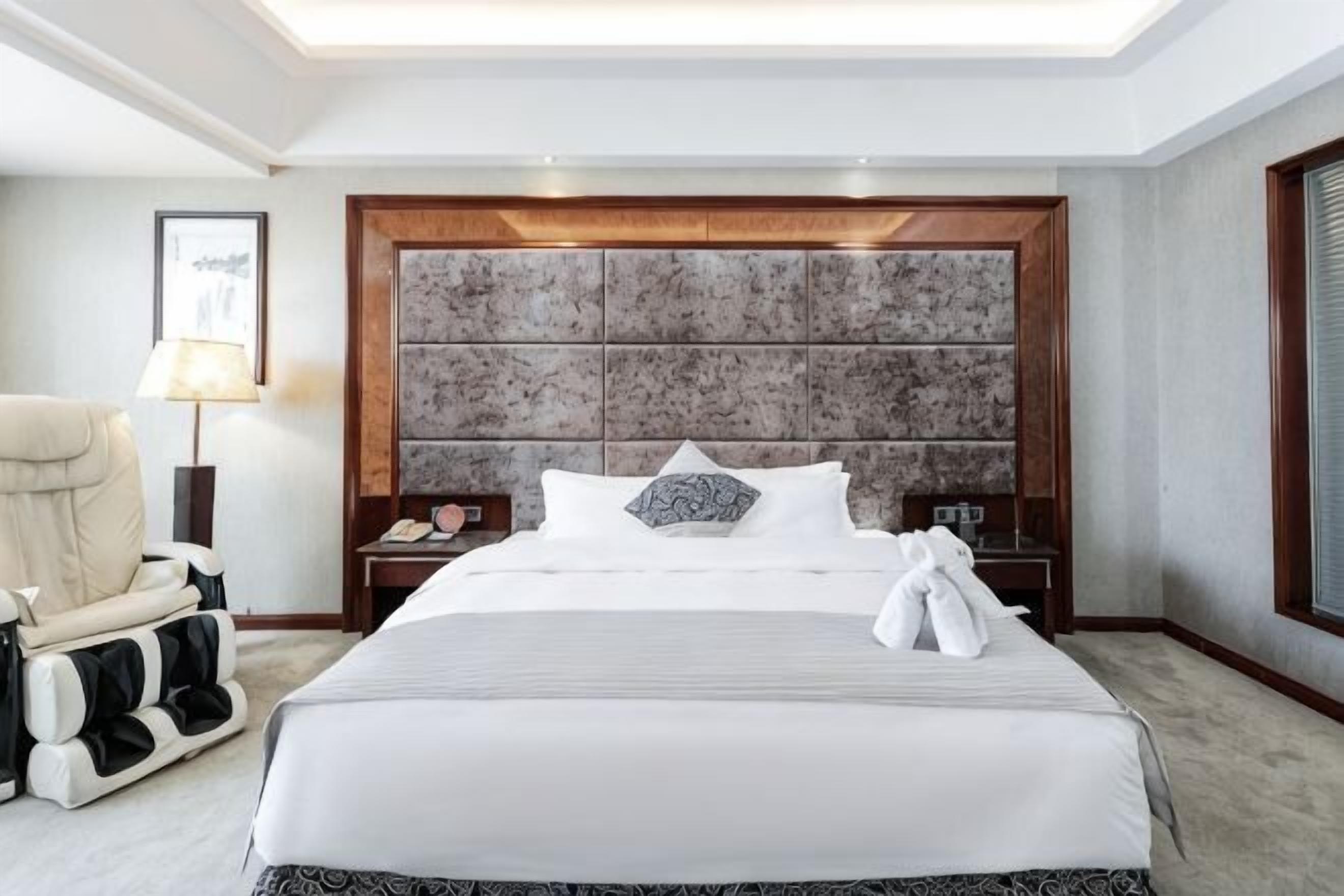 A hotel room with a large bed, a headboard with a textured design, a chair, a nightstand, and a lamp.