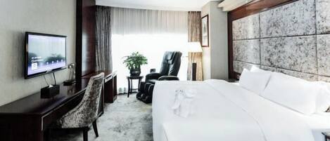 A hotel room with a large bed, a desk with a flat-screen TV, a chair, a potted plant, and a view of the outdoors.