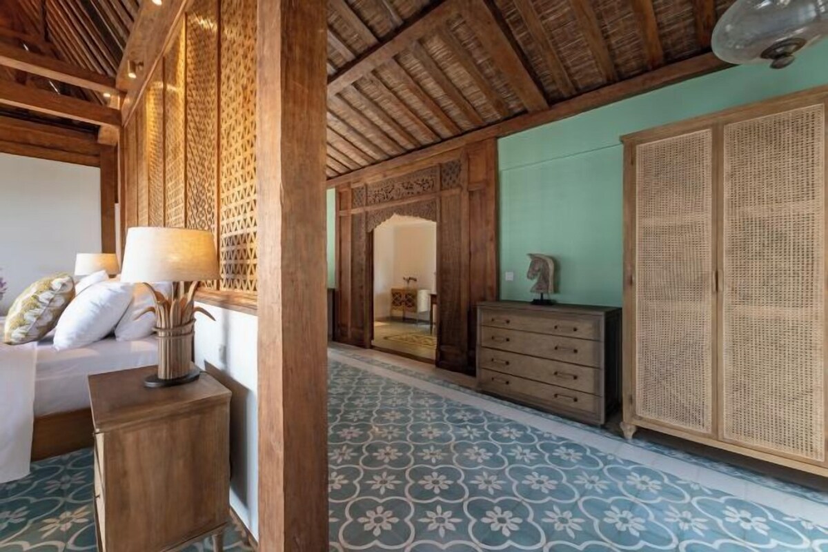 A room with a bed, wooden furniture, a patterned floor, and a traditional woven door.
