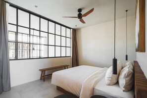 A bedroom with a large window, a ceiling fan, a bed with white bedding, a wooden bench, and a hanging pendant light. - Surf Loft 4 by Alfred in Bali (Cemagi)