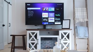 Ein Wohnzimmer mit einem an der Wand montierten Flachbildfernseher, einem Kamin darunter und einem Tisch mit einer Auflage.