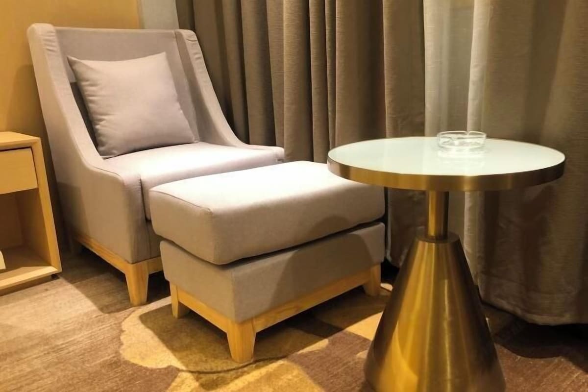 Una habitación de hotel con un sillón gris, un pequeño reposapiés, una mesa redonda con un vaso y una mesita de noche de madera.