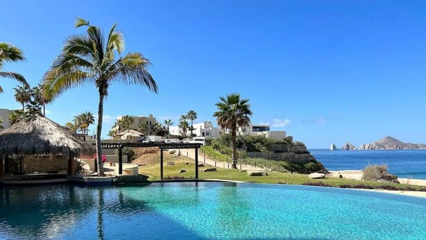 A clear blue pool with a thatched-roof structure and palm trees, overlooking a coastal landscape. - Spectacular Ocean-View  Pet friendly Condo. Walk to Beach, Bars & Dining (Cabo Bello)
