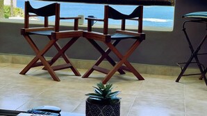 A terrace with two wooden chairs, a small table, a potted plant, and a view of the sea and mountains. - Spectacular Ocean-View  Pet friendly Condo. Walk to Beach, Bars & Dining (Cabo Bello)