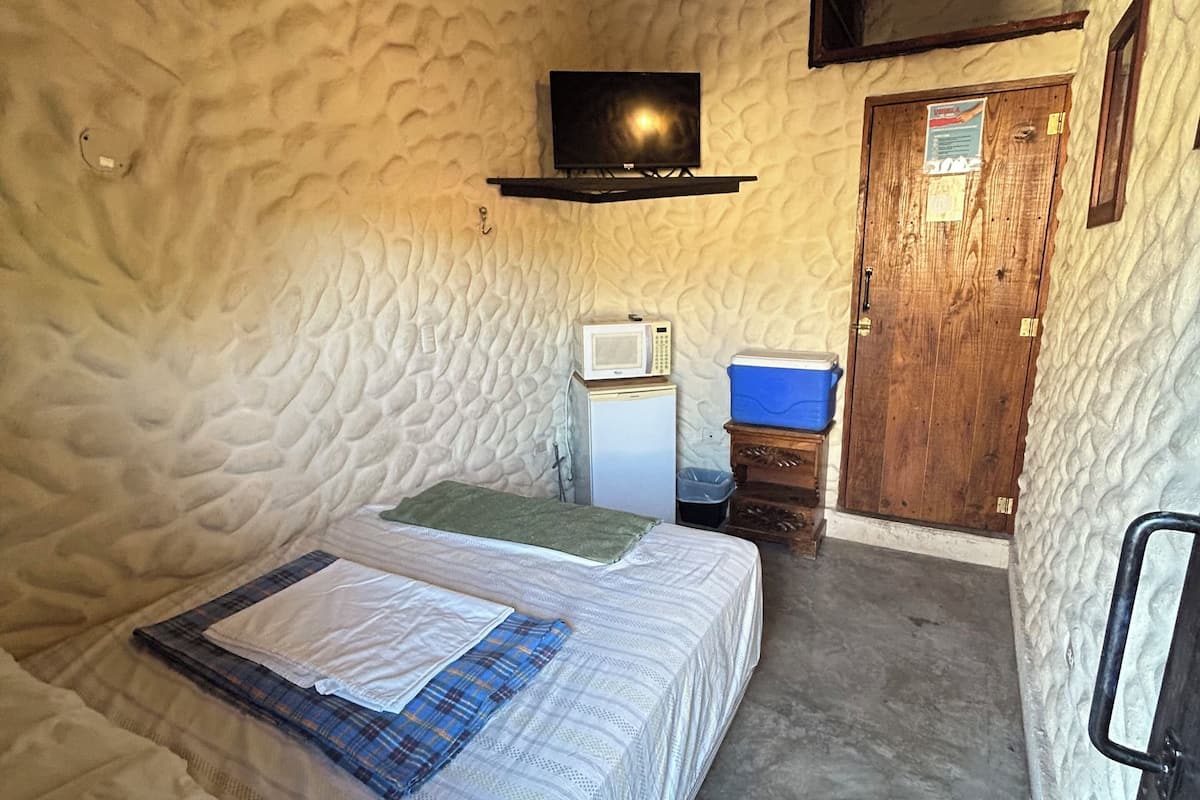 Quarto pequeno e aconchegante com parede texturizada, uma cama com edredom branco e manta xadrez, micro-ondas, mini geladeira, lixeira azul e porta de madeira.
