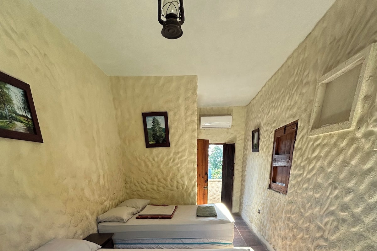 Um corredor estreito leva a duas camas individuais com cabeceiras, uma porta de madeira e uma pequena janela com persianas.