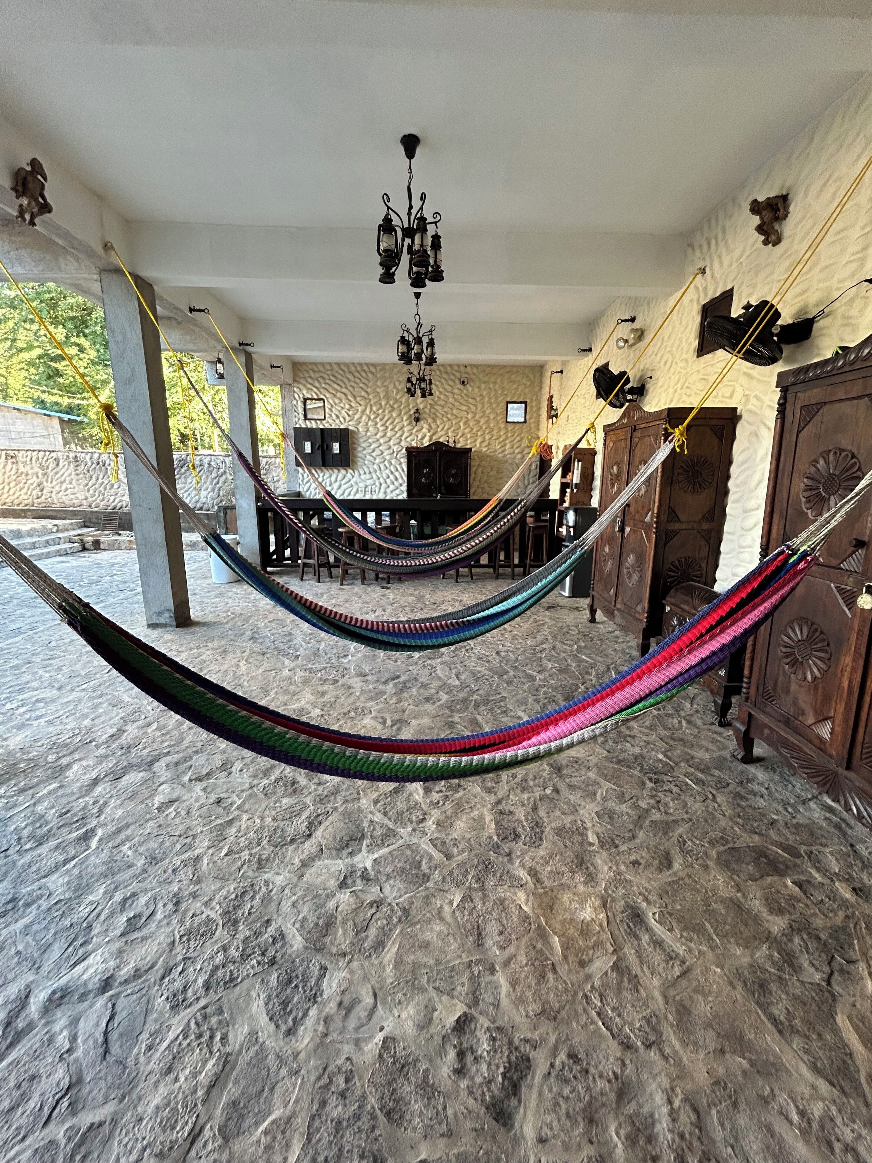 A hammock in a covered outdoor area with a stone floor, a wooden cabinet, and a chandelier.