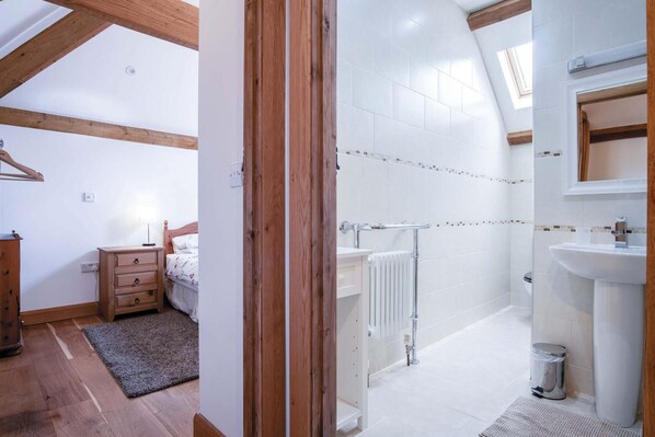 Quarto com cômoda de madeira, mesa de cabeceira e cama. Ao lado, banheiro com chuveiro, vaso sanitário e pia.