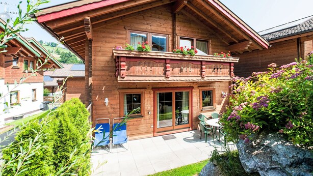 Een houten chalet met een balkon, grote ramen en een terras met buitenmeubels.