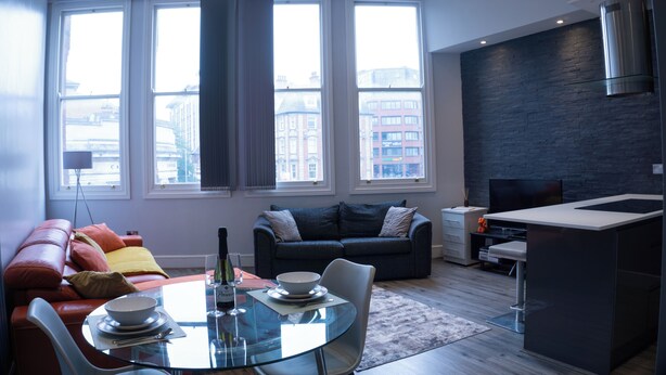 A modern living room with a glass dining table, a sofa, and a kitchen area with a built-in stove. - Spacious Apartment In City Centre - Free Parking (Nottingham)