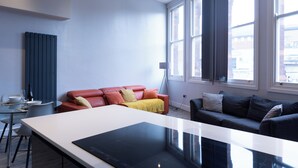 A modern living room with a white table, a red leather sofa, and a dark grey sofa. There are large windows with curtains, a radiator, and a floor lamp. - Spacious Apartment In City Centre - Free Parking (Nottingham)