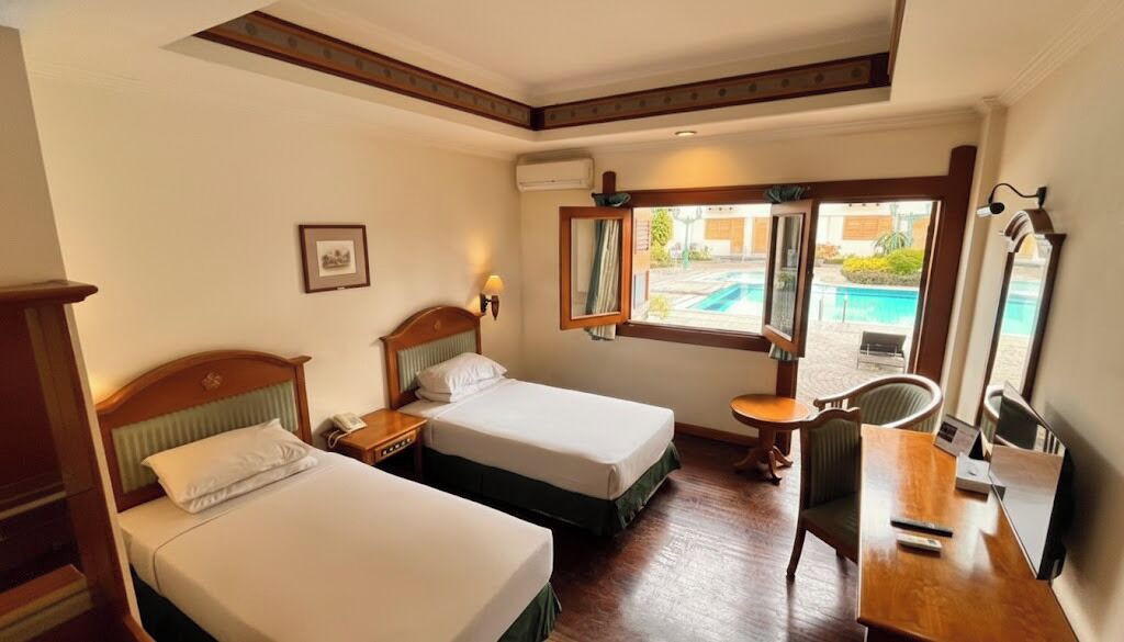 A hotel room with two beds, a desk, a chair, and a window with a view of a swimming pool.