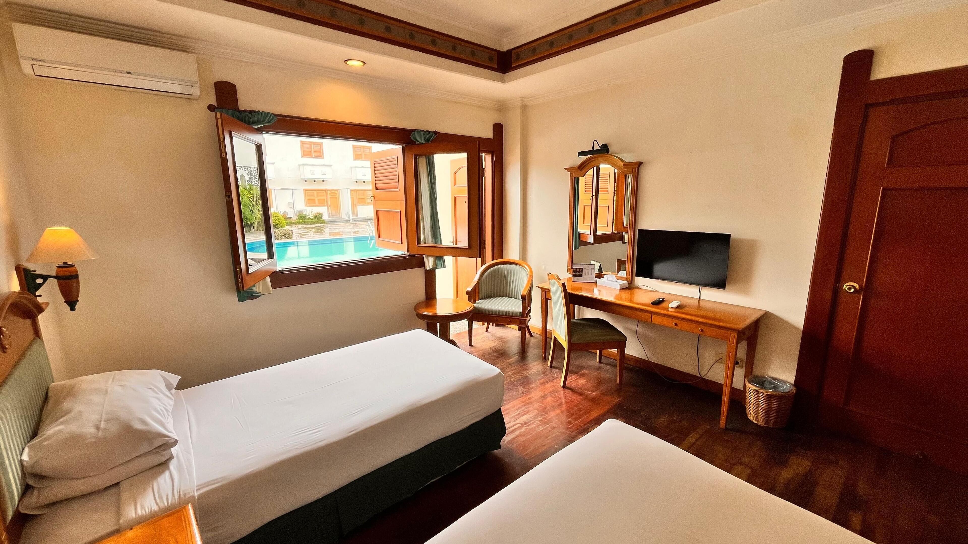 A hotel room with a bed, desk, chair, television, and a window with a view of buildings and a pool.