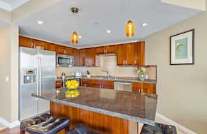A modern kitchen with wooden cabinets, stainless steel appliances, a granite countertop, and pendant lighting. - Maui Resort Rentals: Honua Kai Hokulani - 3 Total Bedrooms, XL Balcony, Lush Courtyard & Beautiful Ocean Views! (Lahaina)
