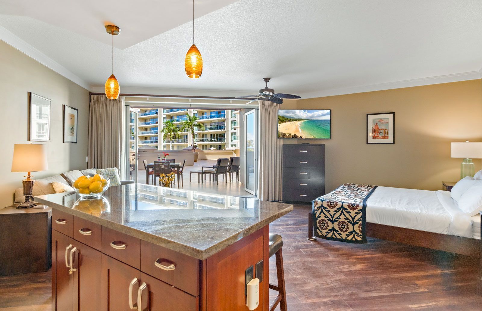 Hotellihuone, jossa on sänky, kirjoituspöytä, jossa on kulho täynnä hedelmiä, ruokailutila, josta on näkymä palmuille, sekä parveke, jolta on näkymä rannalle.