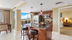 A modern kitchen with a central island, stainless steel appliances, and a dining area with a view. - Maui Resort Rentals: Hokulani 4 Total Bedrooms, Stunning Partial Ocean & Mountain Views + XL Lanai! (Lahaina)