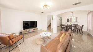 A modern living room with a flat-screen TV, leather sofas, a dining area with a chandelier, and a staircase leading to another level. - Spacious 4BR w Game Room - 15 min to Seaworld (San Antonio)