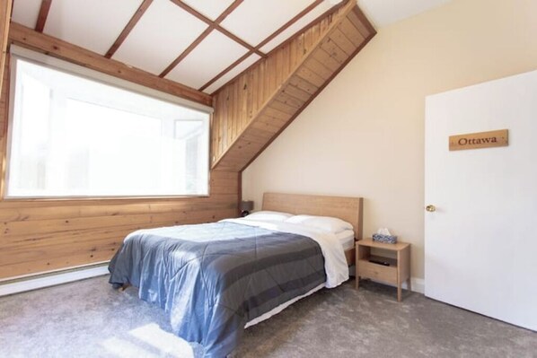 A bedroom with a sloped ceiling, a bed with a blue and gray striped duvet, a bedside table with a lamp, and a door labeled "Ottawa".