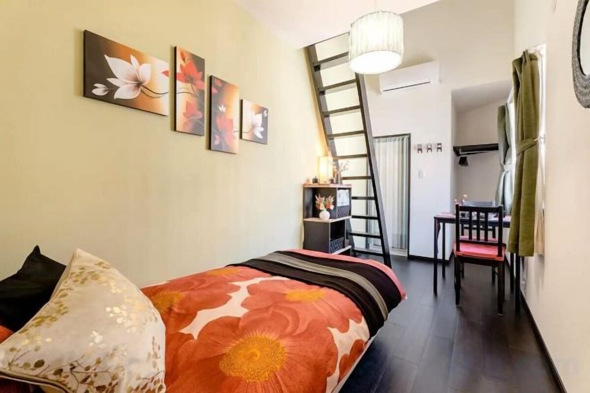 Un dormitorio con cama, almohadas, un escritorio y una silla. Hay obras de arte en la pared y una escalera.