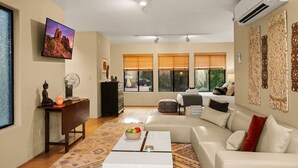 Una sala de estar moderna con un sofá blanco, una mesa de centro con fruta, un televisor en la pared y ventanas grandes con persianas.