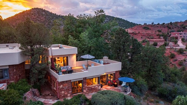 Una casa moderna de dos pisos con un balcón, rodeada de abundante vegetación y un paisaje montañoso.