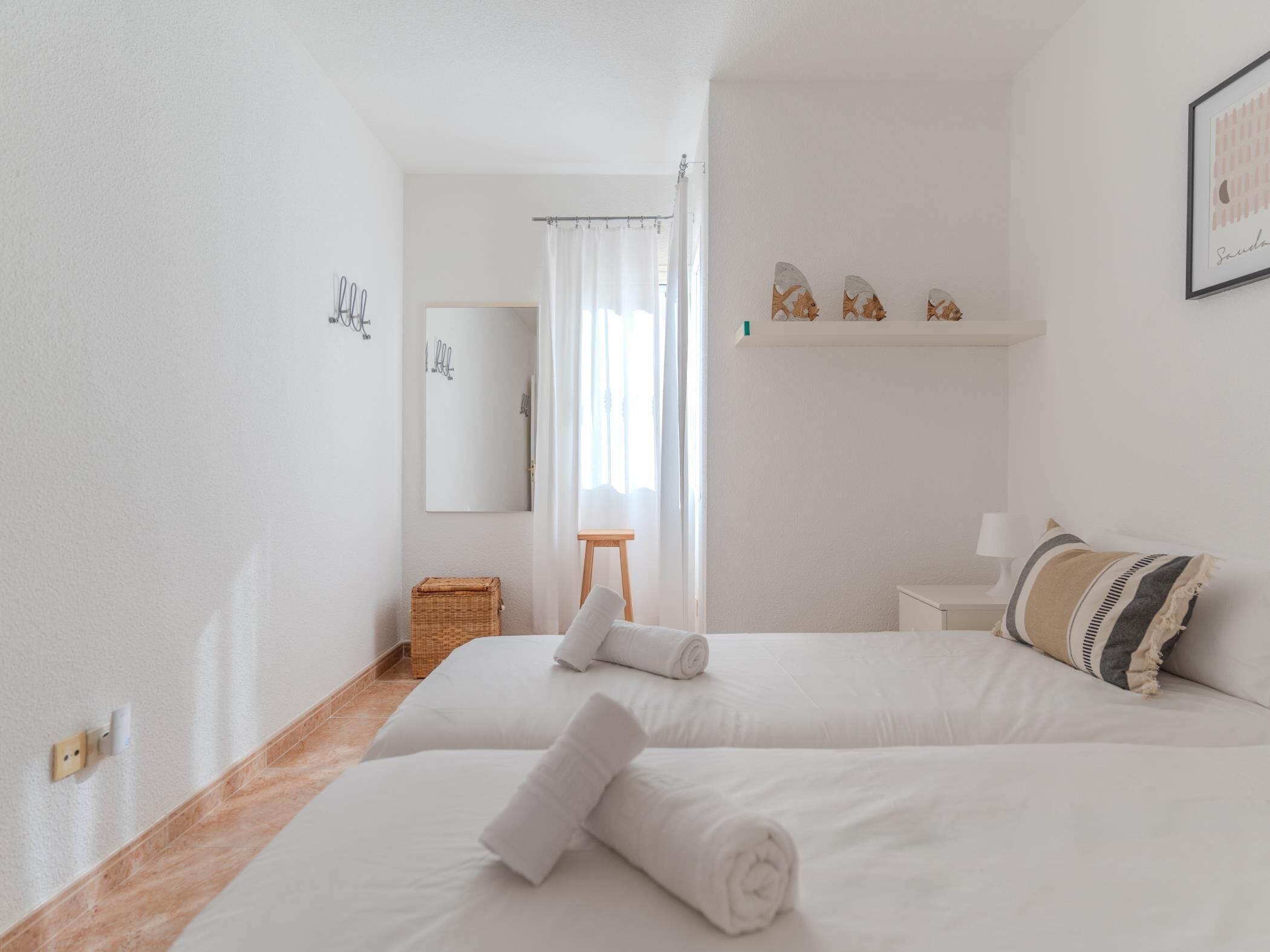 Une chambre d’hôtel avec deux lits, un petit tabouret en bois, un panier en osier, un miroir et une étagère avec des objets décoratifs.