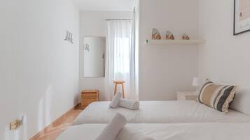 Une chambre d’hôtel avec deux lits, un petit tabouret en bois, un panier en osier, un miroir et une étagère avec des objets décoratifs.
