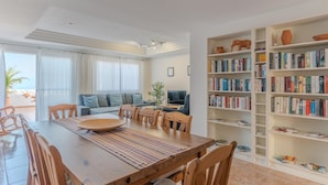 A spacious living area with a wooden dining table, a sofa, and a bookshelf filled with books.