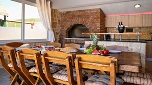 A rustic dining area with a brick oven, wooden chairs, and a table set with food and drinks.