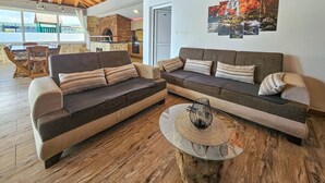 A modern living room with a wooden ceiling, a stone fireplace, a dining area with a wooden table and chairs, and a wall with a clock and nature-themed artwork.
