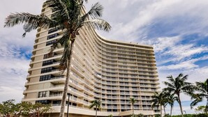 A multi-story hotel with a curved facade, multiple balconies, and palm trees in the foreground. - Modern 2BR Condo l Gorgeous Oceanview (Marco Island)