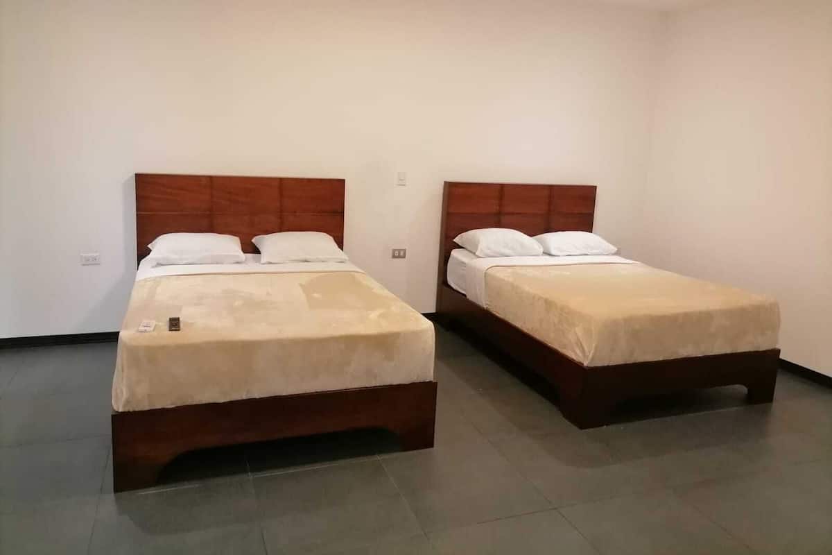 Dos camas individuales con cabeceros de madera en una habitación con paredes blancas lisas y suelo de baldosas.