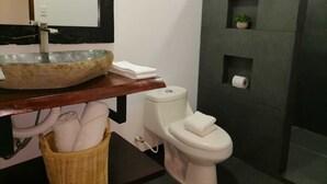 Ein Badezimmer mit einem Waschbecken aus Stein, einer Toilette, einem Korbwannenaufsatz und einer kleinen Pflanze an einer dunklen Wand.