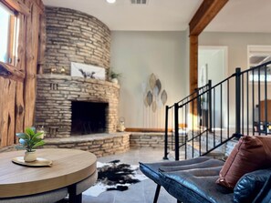 Une cheminée en pierre, une table en bois ornée d’une plante, un canapé en cuir et un escalier avec une rampe noire.