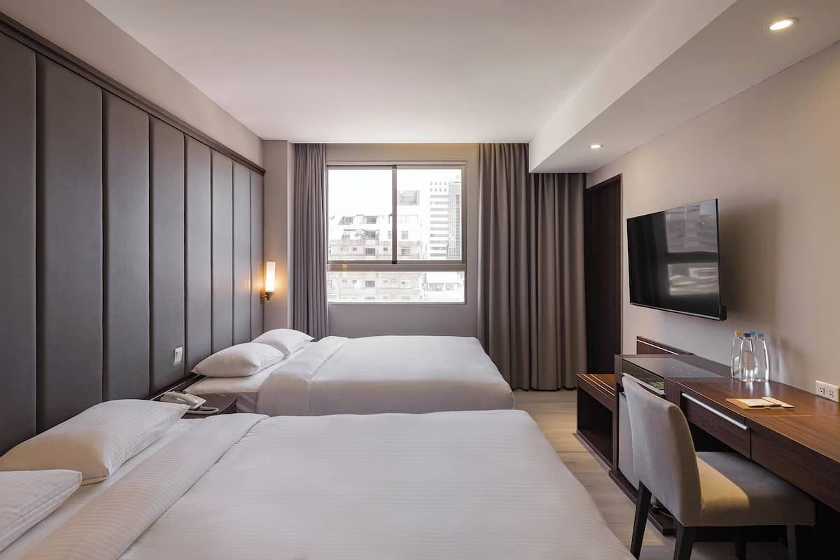 A hotel room with two beds, a desk, a chair, a TV, and a window with a city view.
