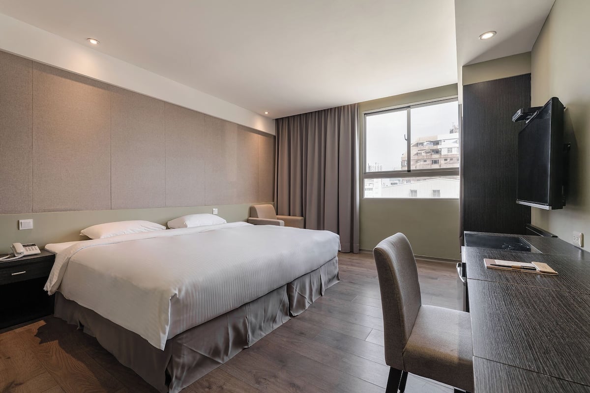 A modern hotel room with a large bed, a desk with a chair, a flat-screen TV, and a city view through the window.