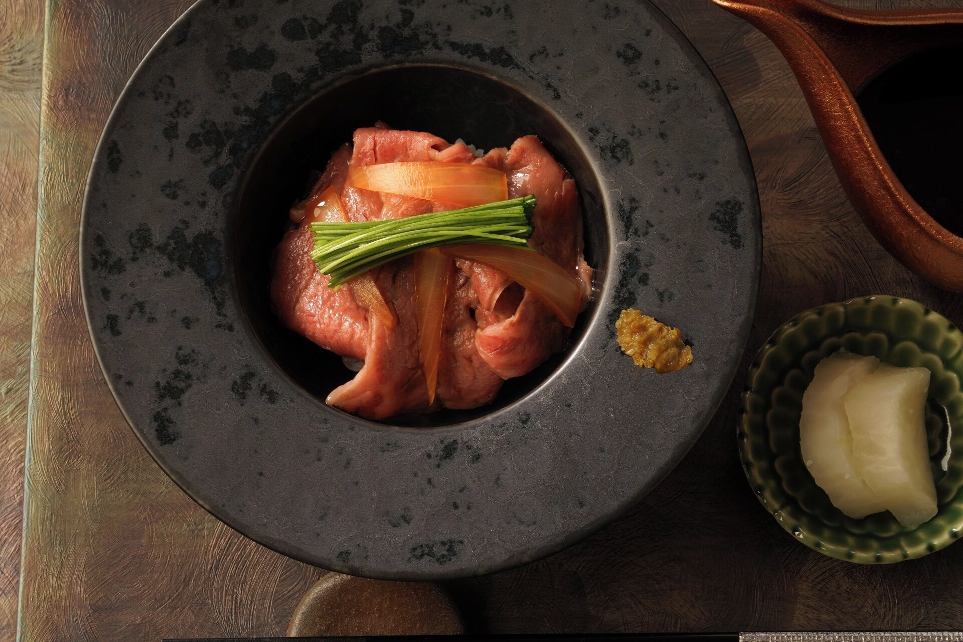 A bowl of raw meat with onions and herbs, a small bowl of rice, and a clay pot on a wooden surface.