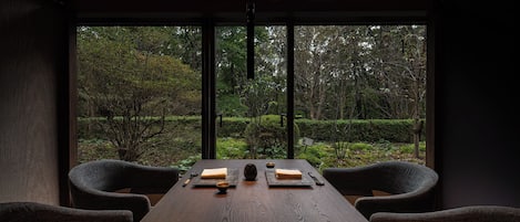 A dining table with two chairs, a bowl, chopsticks, and a small plate in a room with large windows overlooking a forest.