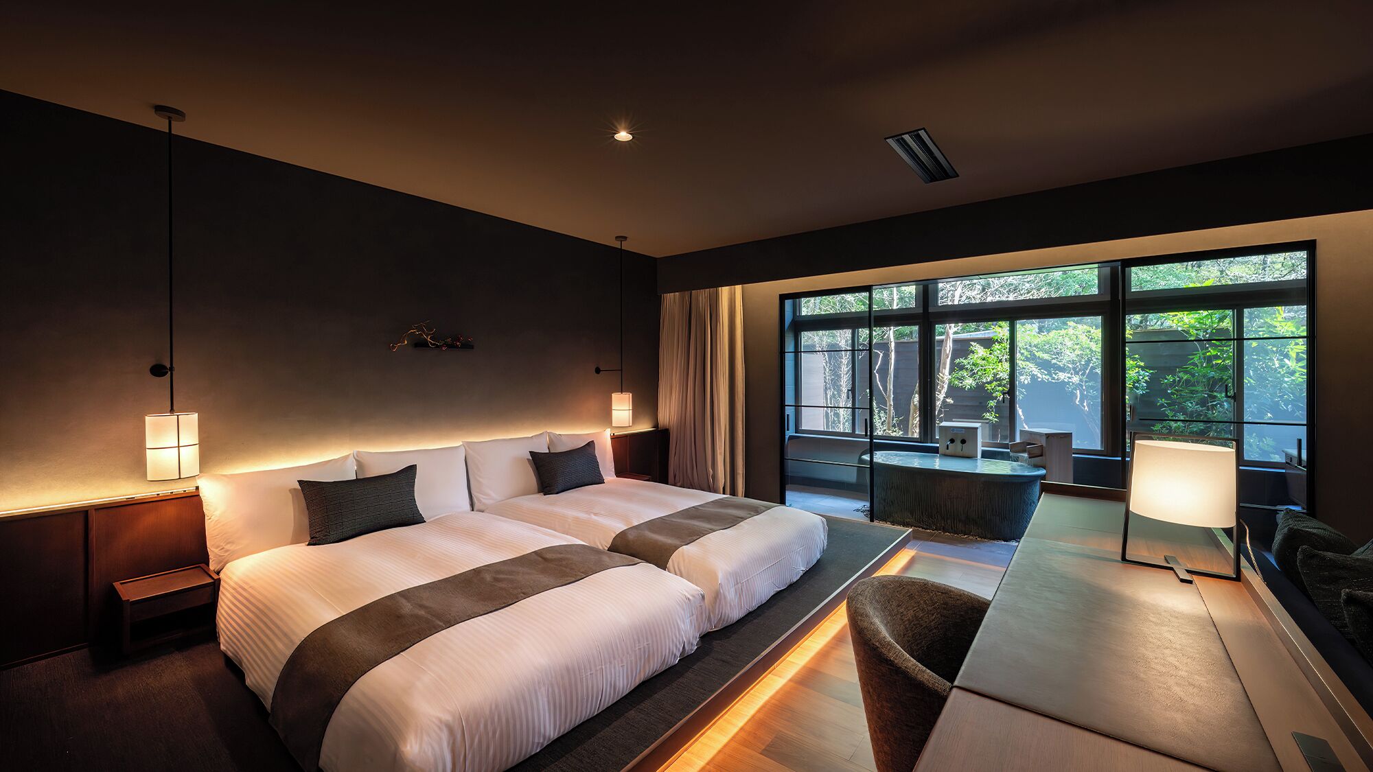 A modern hotel room with a large bed, bedside lamps, a desk, and a view of greenery through large windows.