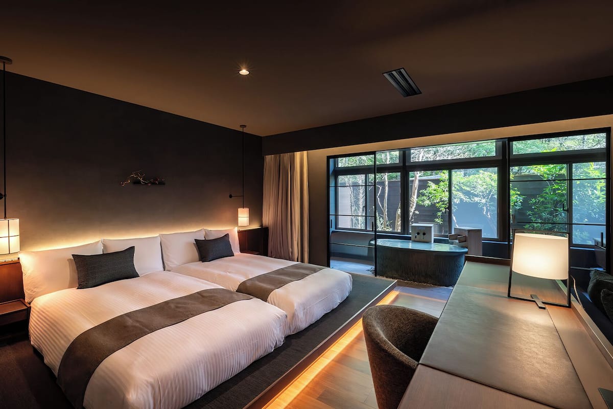 A modern hotel room with a large bed, bedside lamps, a desk, and a view of greenery through large windows.