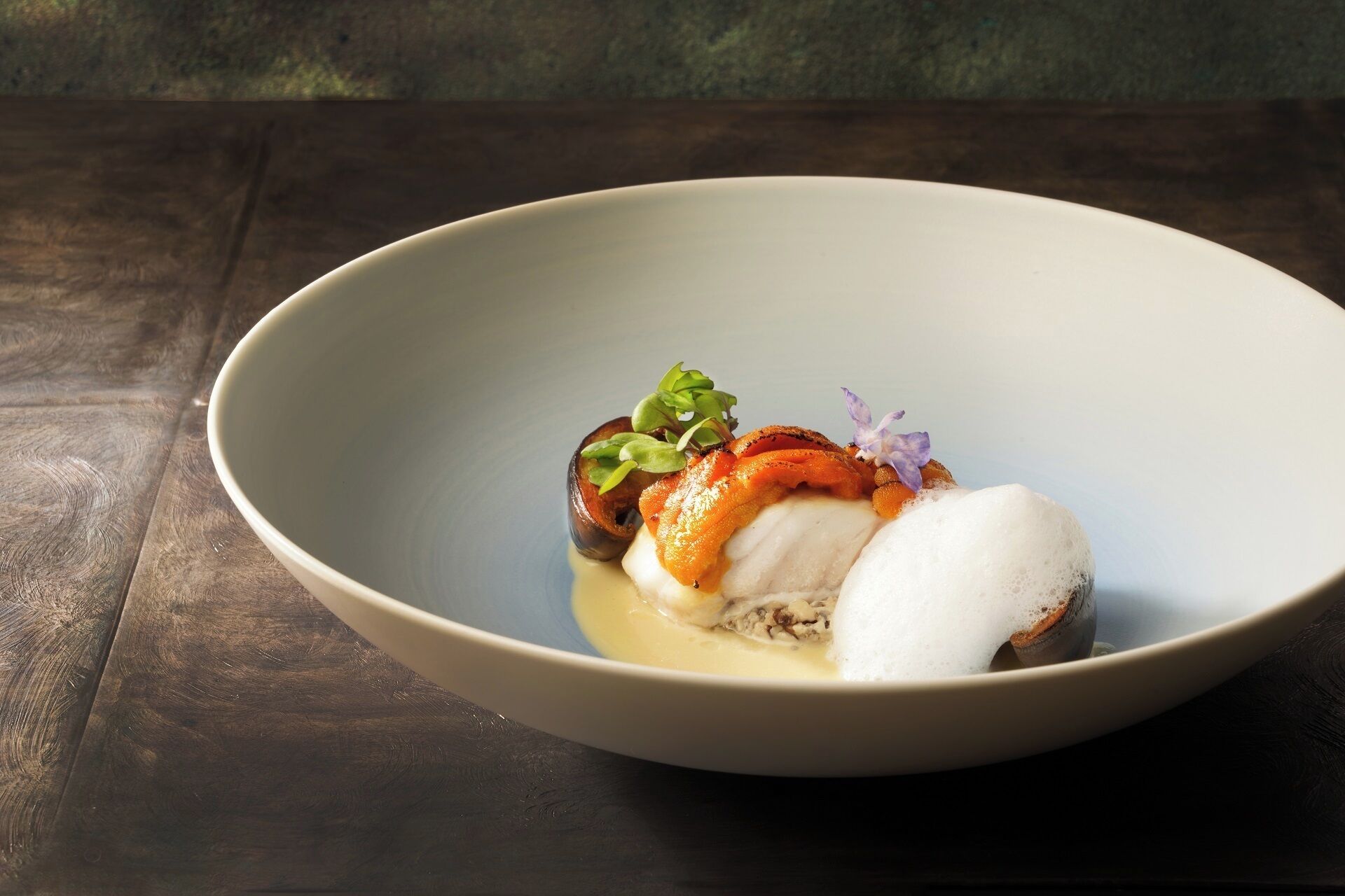 A bowl of food with a creamy sauce, a piece of fish, and some greens on a wooden table.
