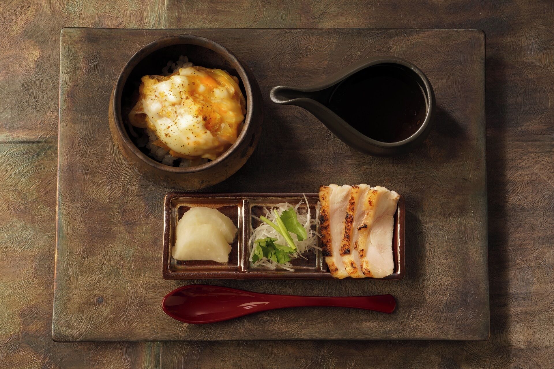 A meal consisting of a bowl of rice topped with a poached egg, a small dish with pickled vegetables, and grilled fish, served on a wooden tray with a red spoon.