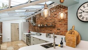 A modern kitchen with a brick wall, a large clock, and a wooden cutting board. - Lodge Farm - Stunning 3 BD with Garden (Oxford)