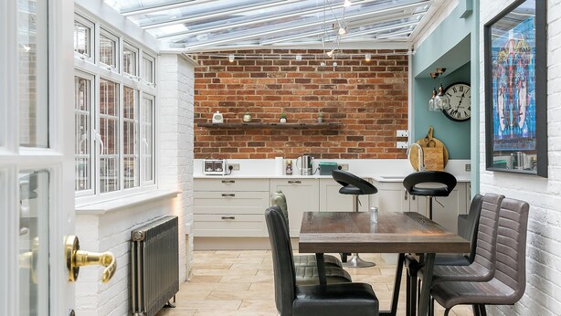 A compact kitchen with white cabinetry, a brick wall, and a dining area with a wooden table and chairs. - Lodge Farm - Stunning 3 BD with Garden (Oxford)