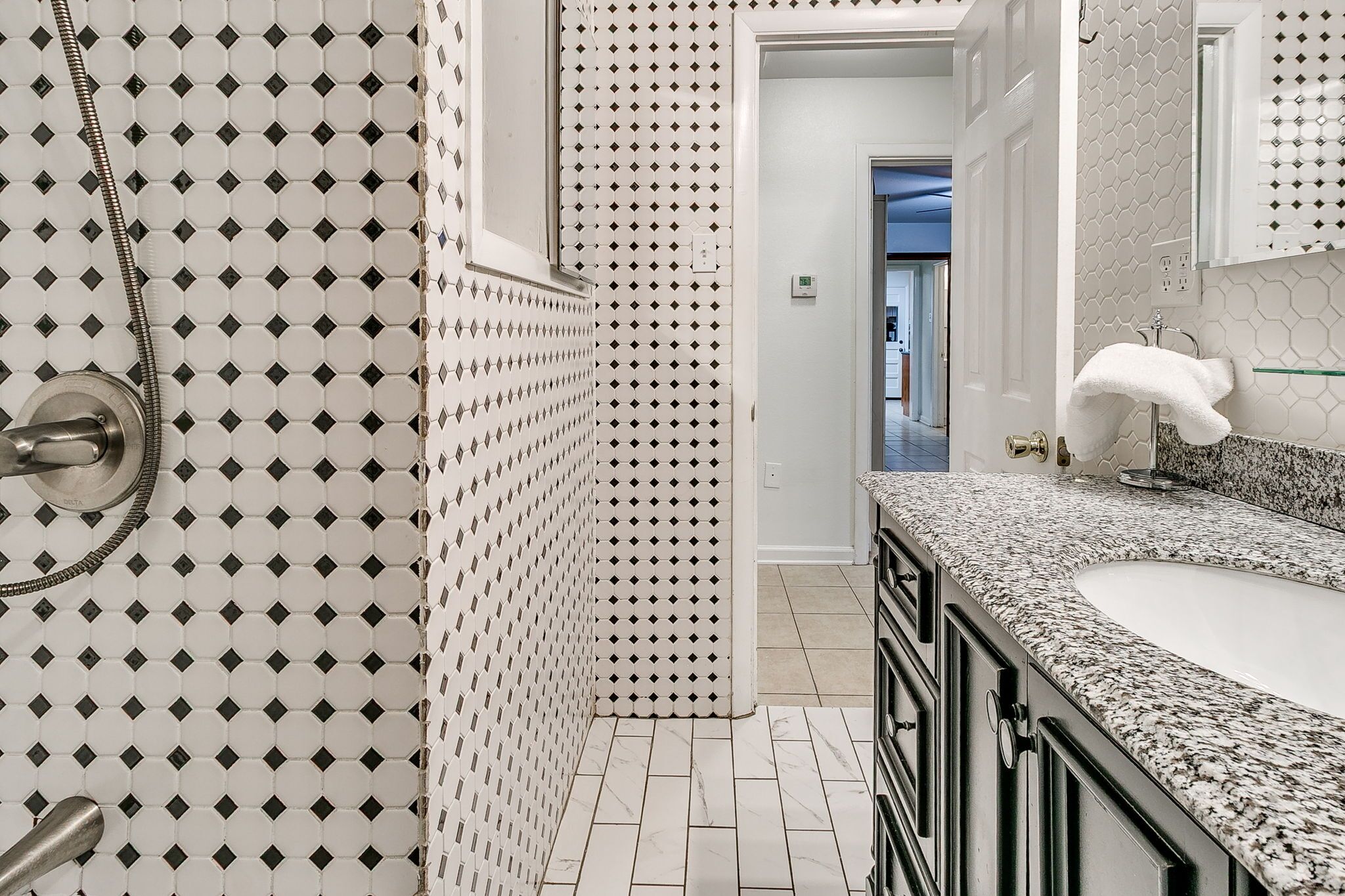 A bathroom with a white and black patterned tile floor, a granite countertop, and a white sink with a glass shelf.