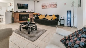 A living room with a fireplace, a black sofa with yellow cushions, a coffee table, and a TV mounted on the wall.