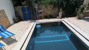 A rectangular swimming pool with clear blue water, surrounded by stone tiles and a few lounge chairs.