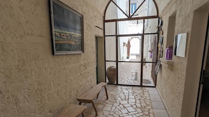 A corridor with a stone floor, a wooden bench, a framed painting, and a view through a glass door.