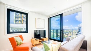 A modern living room with a TV, orange and grey sofas, a small coffee table, and large windows offering a city view.